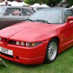 El Alfa Romeo SZ, conocido también como “Il Mostro”, es uno de los deportivos más polémicos jamás construidos por la marca italiana. Diseñado por Zagato y con la firma de Robert Opron, combinaba un aspecto radical con un motor Busso V6 de 210 CV. Su chasis derivaba del Alfa 75 y gran parte de sus componentes venían de modelos ya existentes, lo que contrastaba con su aspecto futurista. Apenas se fabricaron unas 1.000 unidades, lo que lo convierte en una rara joya para coleccionistas.